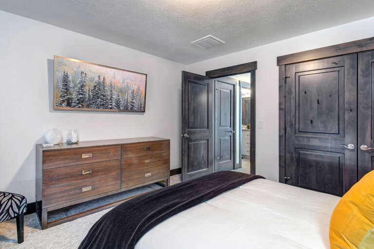 Settle into your bedroom featuring a cozy bed, polished wood dresser, and peaceful mountain art. Dark-stained doors lead to closet and en-suite, offering all the comfort you need.