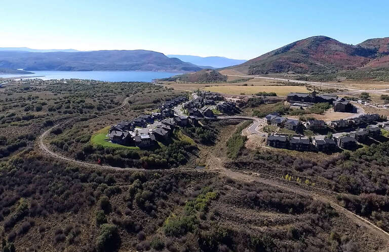 Aerial view captures the property nestled in a scenic valley with mountain ranges and a tranquil lake visible in the distance, surrounded by natural landscape.
