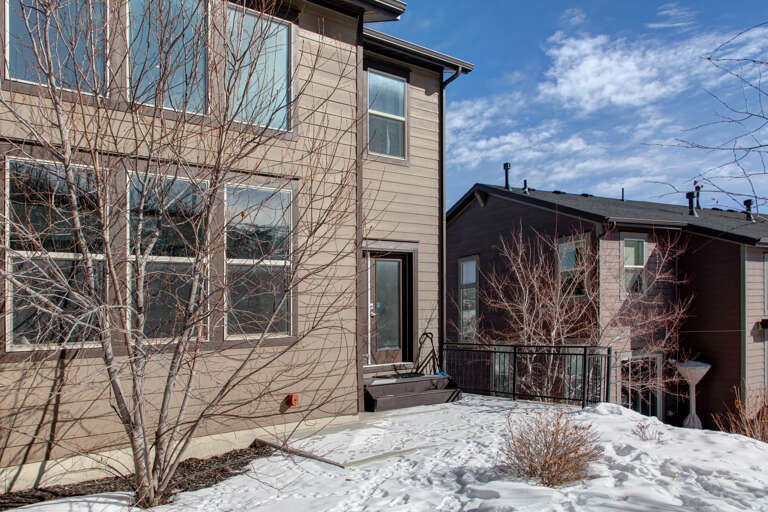A contemporary property set in a quiet winter neighborhood, featuring multi-level architecture with modern siding, large windows, and nearby mountain views beneath a bright blue sky.