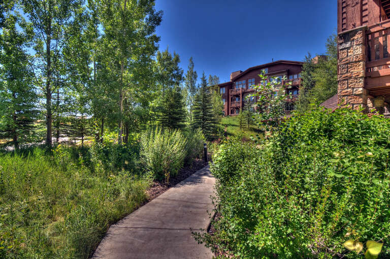 A winding path leads through lush landscaping and mature trees toward the property's distinctive architecture, blending natural beauty with mountain resort charm.
