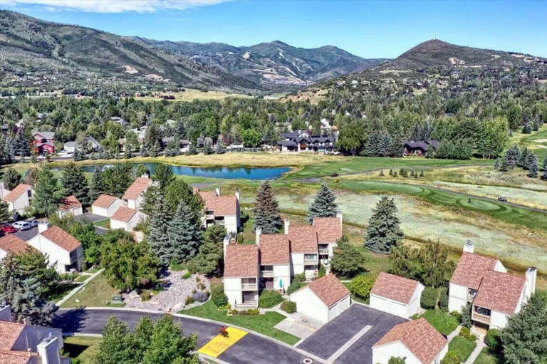 This aerial view showcases the property nestled in a scenic mountain valley, surrounded by evergreen forests, rolling hills, and a tranquil pond—offering a peaceful natural setting for your getaway.
