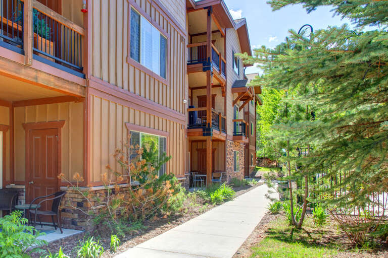 A multi-level vacation rental building framed by lush evergreens and landscaped gardens, offering a peaceful mountain-area retreat with private balconies and ground-floor patios.