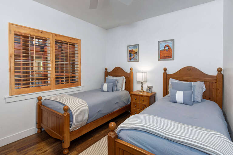 Your peaceful twin bedroom features cozy wood-framed beds with soft blue bedding, warm plantation shutters, and charming national park artwork—ideal for restful nights after exploring the area.