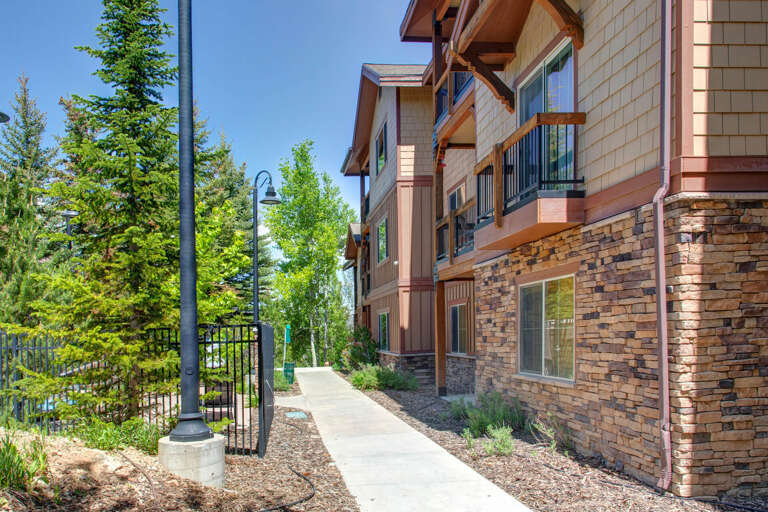 Multi-level vacation building with stone and wood siding nestled among tall evergreens. A welcoming pathway and mountain-style architecture create a warm arrival experience in this scenic natural setting.