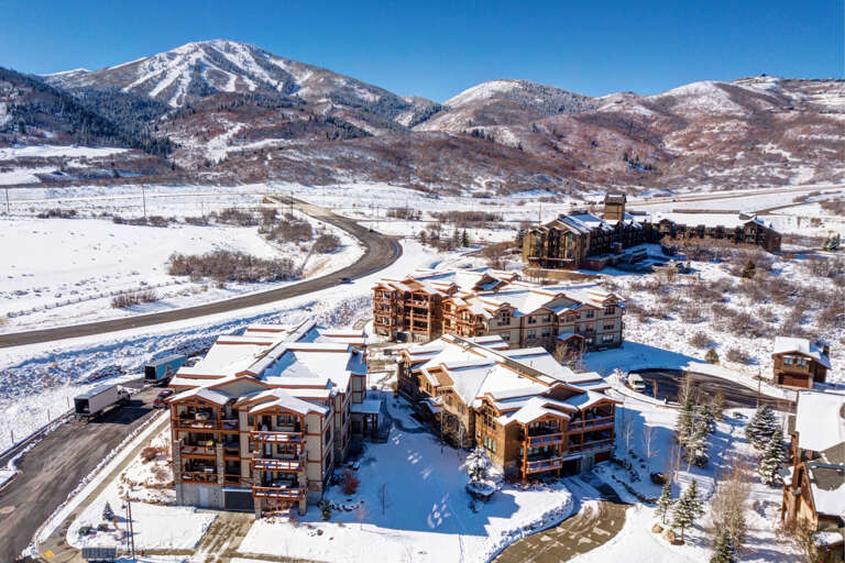 An expansive winter resort nestled in a pristine mountain valley, featuring multiple lodges amid snowy terrain with ski slopes rising in the background. The property sits along a scenic mountain road surrounded by natural landscape.