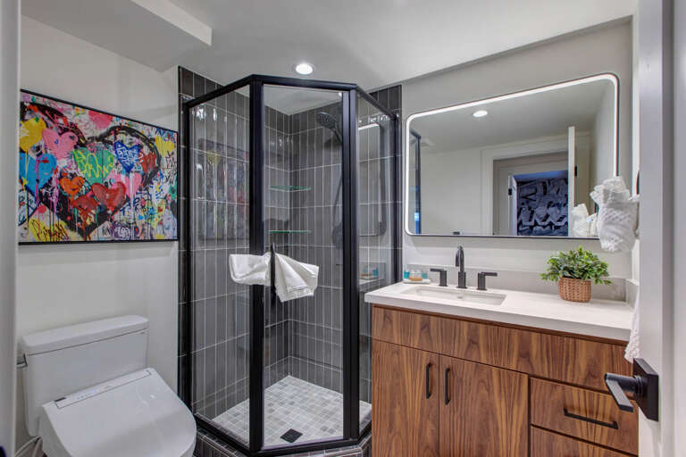 Your private bathroom blends modern style with comfort—featuring a sleek glass shower, warm wood vanity, and a bright backlit mirror to start your mornings refreshed.
