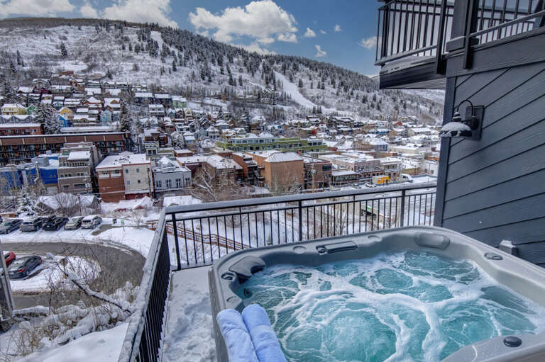 A private hot tub on a snowy balcony overlooks a picturesque winter mountain town with ski slopes visible on the hillside, offering a serene alpine escape.