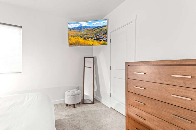 Relax in your serene bedroom with a wall-mounted TV showcasing stunning mountain views. Soft natural light and modern furnishings create a peaceful retreat for restful evenings.