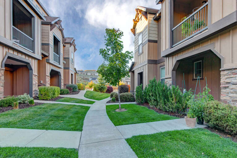 Multi-unit residential complex featuring manicured lawns, winding pathways, and mature landscaping with scenic mountain backdrop under dramatic cloudy skies.