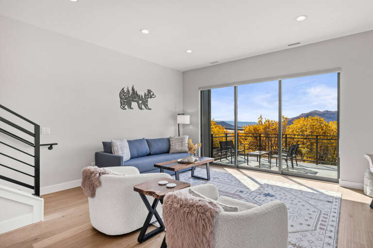 Relax in your bright, modern living room with mountain and lake views beyond the private balcony—a peaceful retreat framed by golden fall foliage.