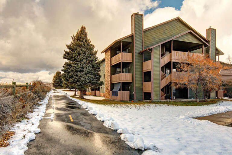 A green multi-story building with wooden balconies and stone accents sits along a snow-dusted path, surrounded by evergreen trees and mountain views, marking the property entrance.