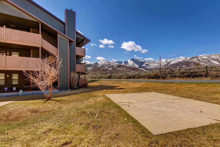 Multi-level vacation rental property nestled in scenic mountain landscape with expansive lawn, private balconies, and dramatic snow-capped peaks under clear blue skies.