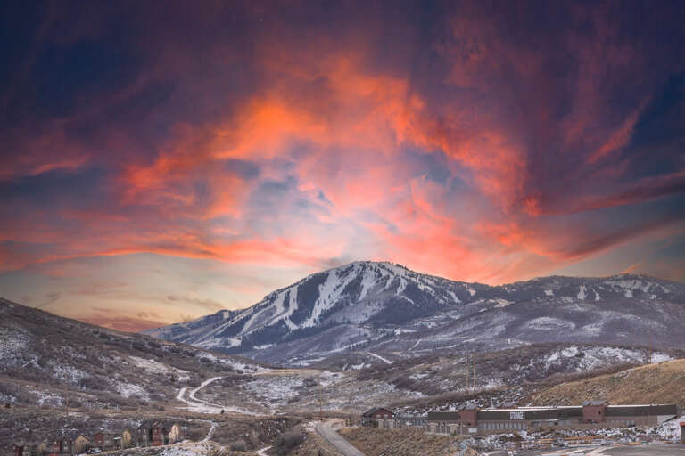 Dramatic sunset paints the winter sky in brilliant oranges and purples above snow-covered mountain slopes and valley community.