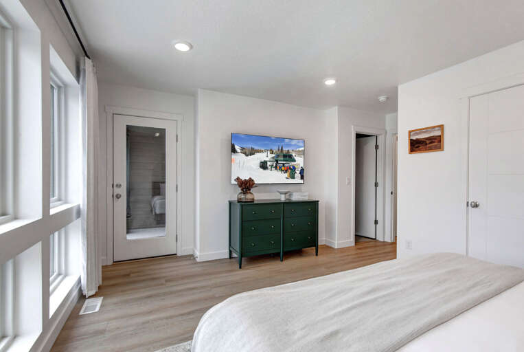 Relax in your spacious bedroom featuring a smart TV, elegant green dresser, and glass door leading to your private ensuite bathroom.