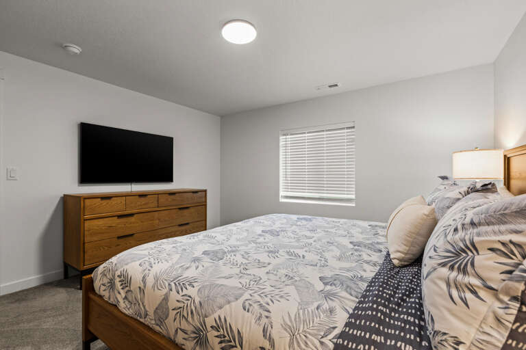 Unwind in your cozy bedroom with warm wood accents, a plush bed with tropical-inspired linens, and a wall-mounted TV for your favorite shows. Natural light filters in through window blinds.