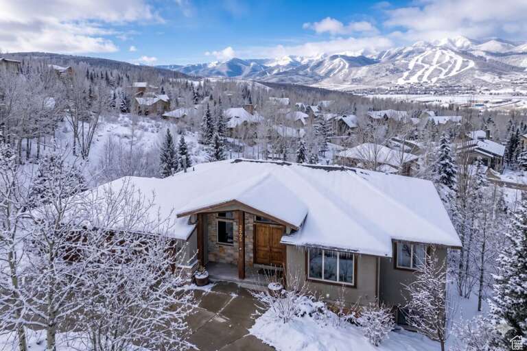 Aerial view of a snow-covered mountain home nestled in a forested neighborhood, with ski slopes and pristine winter peaks forming the dramatic backdrop.