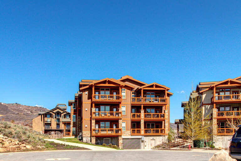 Rustic mountain architecture featuring natural wood accents and stone details, set against a scenic hillside backdrop. The multi-level buildings showcase spacious balconies with breathtaking views.