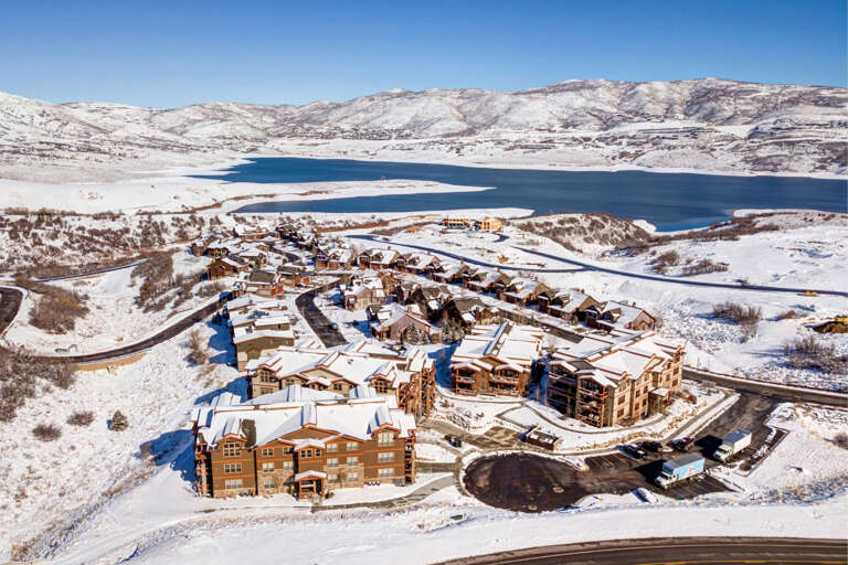 Aerial view of a snow-covered resort community nestled beside a stunning alpine reservoir, surrounded by dramatic winter mountain terrain and pristine wilderness.