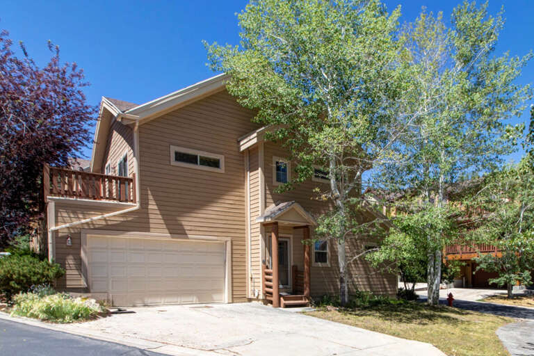 A welcoming two-story vacation home with a spacious garage and private balcony, nestled among tall aspens and mountain greenery under a brilliant blue sky.