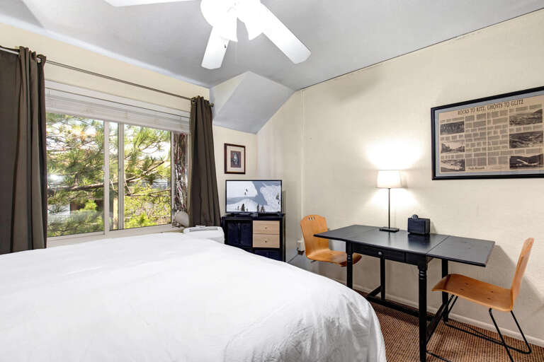 Your bright bedroom invites peaceful mornings with vaulted ceilings, crisp linens, and a workspace bathed in natural light filtering through tall windows overlooking lush greenery.