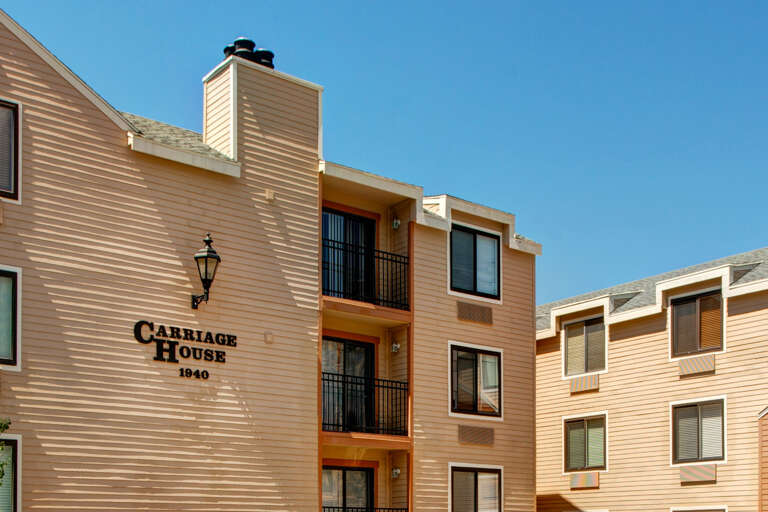Carriage House building features warm siding, private balconies, and a charming exterior lamp under clear blue skies.