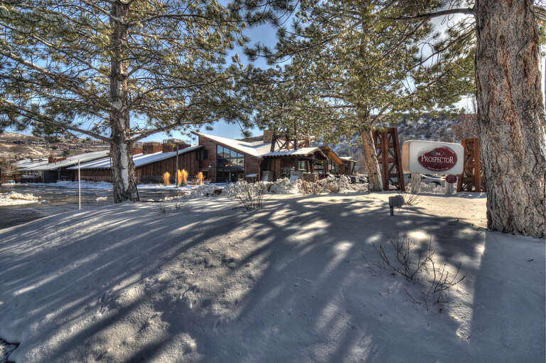 Snow-covered grounds lead to a rustic mountain lodge surrounded by tall pines, offering a peaceful winter retreat beneath brilliant blue skies.