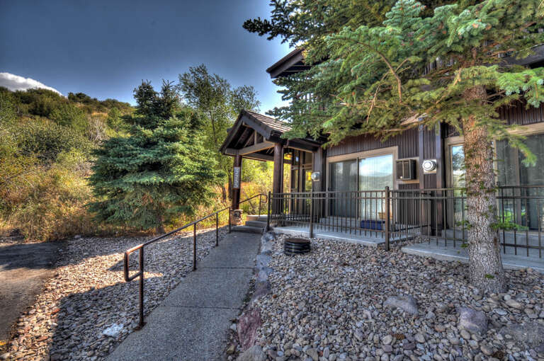 A charming cabin entrance framed by towering pines, surrounded by natural landscaping and accessible pathways leading to the welcoming front door.