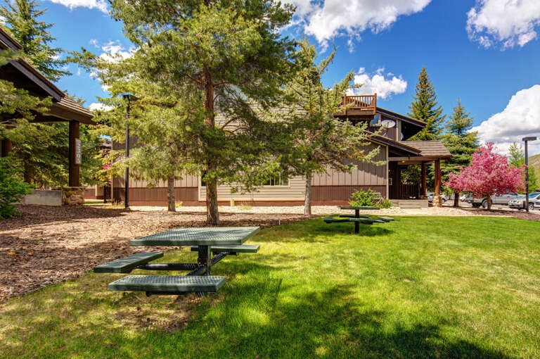 The property features well-maintained communal green spaces with picnic areas, framed by mature evergreen trees and colorful flowering landscaping, set against a backdrop of mountain resort architecture.