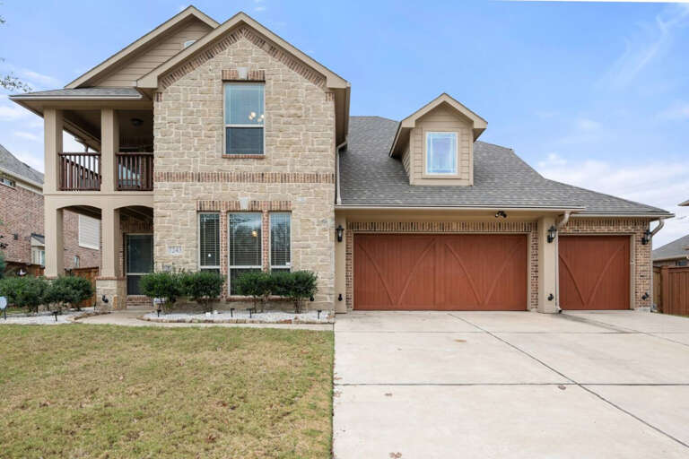 View from street of front of house, showing 3 car garage (available to guests), large driveway, front entryway, and 2nd floor balcony.