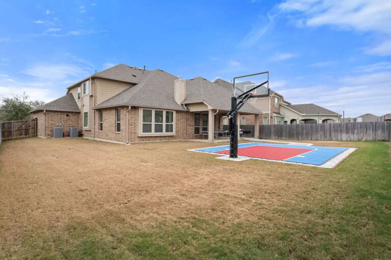 Full backyard view showing side gate and half court basketball court in backyard to enjoy with basketballs provided, as well as patio with ping pong, and extra grass area for added activities.