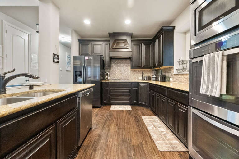 Downstairs full kitchen view from breakfast nook, showing island sink and countertop prep areas, microwave, double oven, looking back toward fridge and stove area with various countertop appliances.