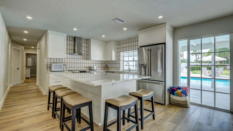 Everything you need to cook a real meal — double oven, dishwasher, stainless fridge, and a large island with bar seating. The kitchen looks out to the pool through the glass door.