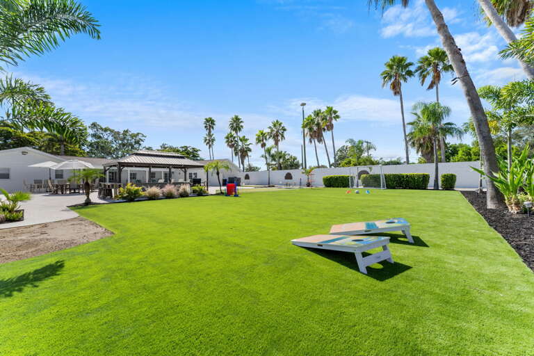 Your full backyard under open sky — cornhole on the turf, gazebo at the edge, and palms lining the fence. More room than your whole street back home.
