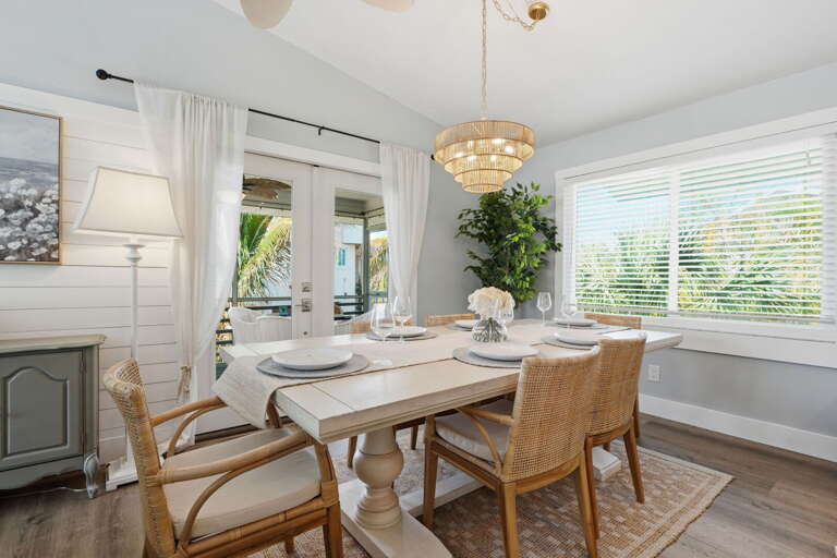 Gather around the white farmhouse table for dinner — rattan chairs, a gold chandelier above, and shiplap wall behind you.