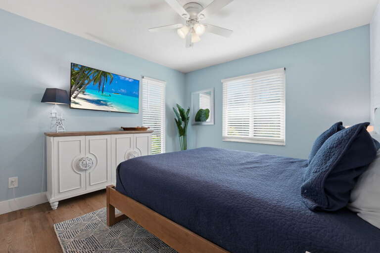 Settle into the queen bedroom — navy bedding, white dresser, mounted Smart TV, and two windows filling the room with light.