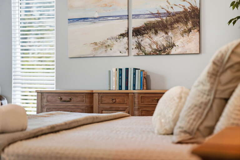 Wake up to coastal art above the dresser — books stacked on the shelf and neutral pillows keeping the master bedroom calm.