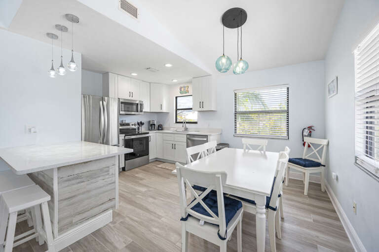 Set a real table two steps from the stove — white cabinets top to bottom, stainless appliances, dining chairs already pulled out.