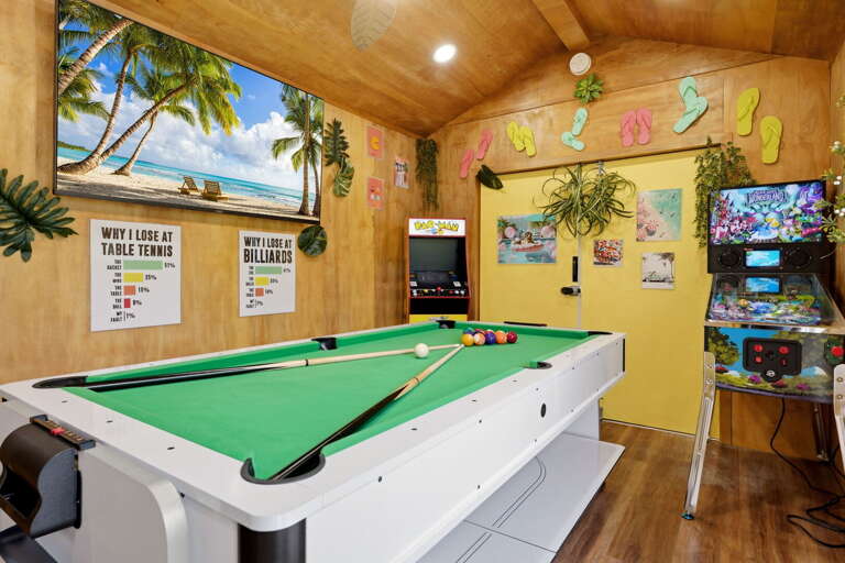 The full room: pool table in the center, PAC-MAN on the left, pinball on the right, and a beach scene playing on the wall above it all.