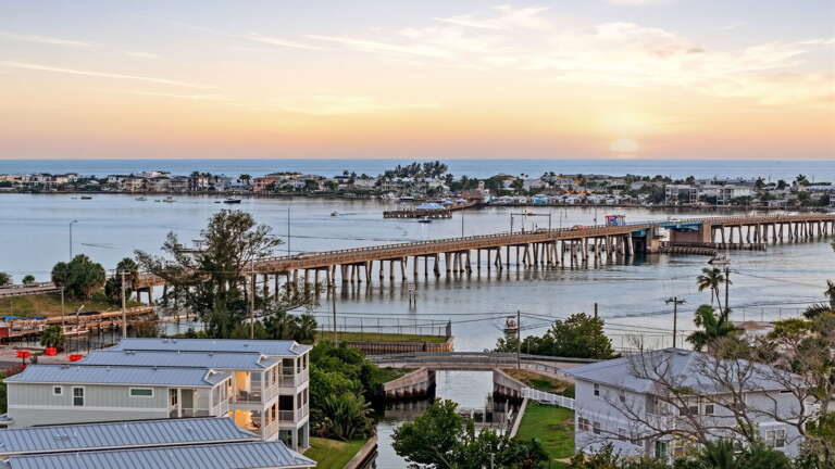 Blue-sky aerial with open water views in every direction - the home anchors the foreground as Sarasota Bay stretches broadly to Anna Maria Island beyond.