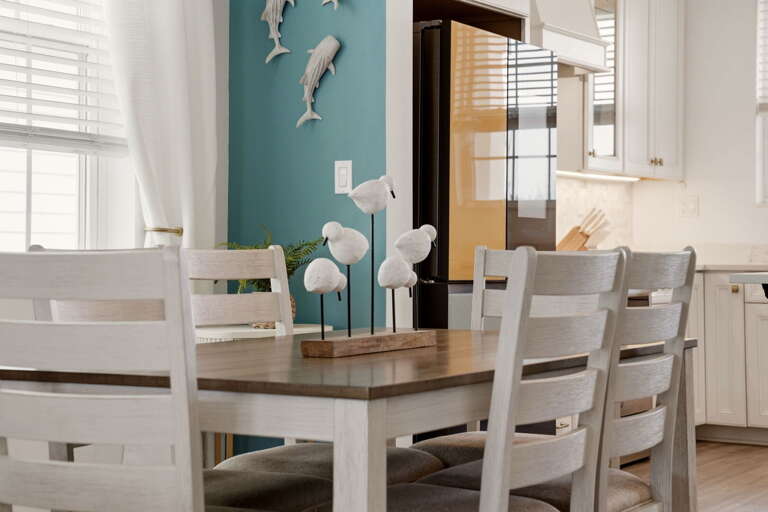 Dining room detail featuring the wood-topped table with nautical shorebird decor and a peek into the bright, fully equipped kitchen beyond.
