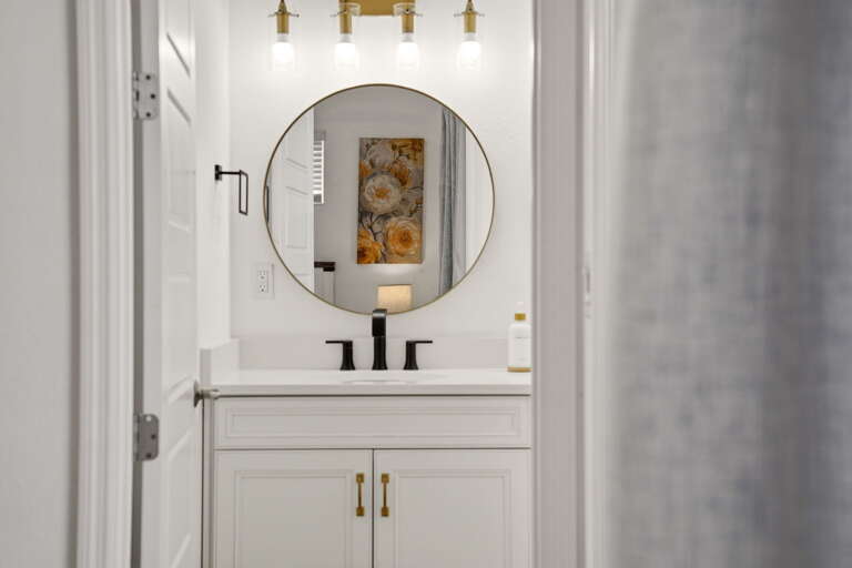 Primary bath vanity detail: a single gold-framed round mirror, gold globe vanity light, and matte black fixtures reflect the bedroom's warm tones and floral artwork beyond.