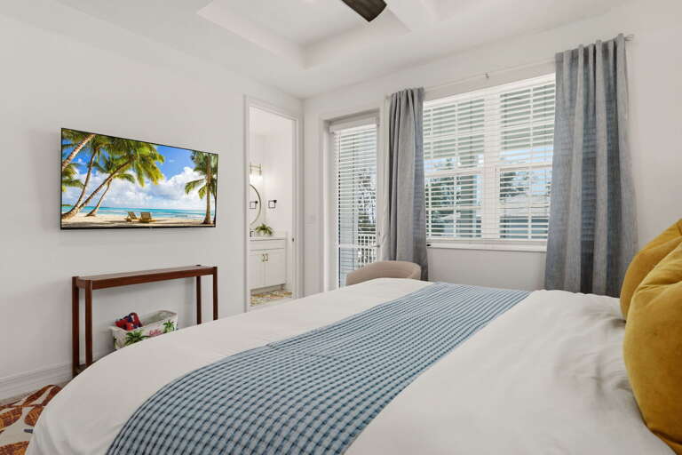 The primary bedroom from the foot of the bed, showing the wall-mounted TV, balcony door, and the entrance to the spa-style ensuite bath.