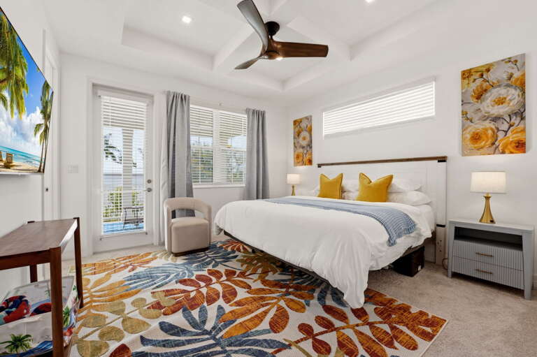 The primary suite is a true retreat - a king bed with mustard yellow pillows, tray ceiling with ceiling fan, wall-mounted TV, private balcony, and a vibrant tropical area rug.