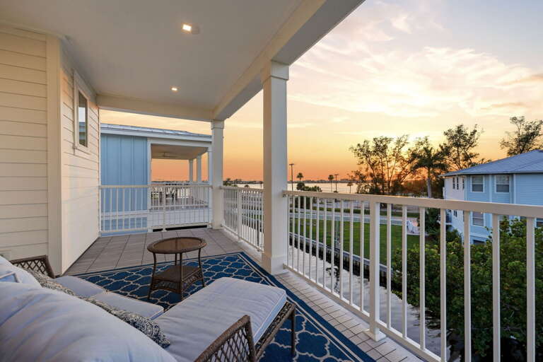 The rear balcony lounge is pure Florida living - a wicker loveseat creates an intimate retreat, with the waterway and bay glowing golden in the evening sky beyond the white railing.