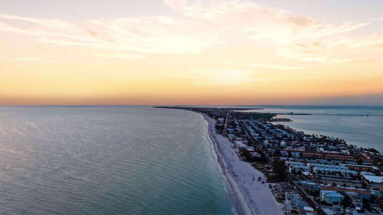 A sweeping aerial along Anna Maria Island's coastline at sunset - miles of pristine white sand, turquoise Gulf waters, and beach-community homes