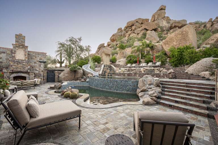 Experience a private desert oasis featuring a stunning multi-level pool, waterslide, outdoor fireplace, and luxurious lounge areas, all surrounded by breathtaking natural rock formations.