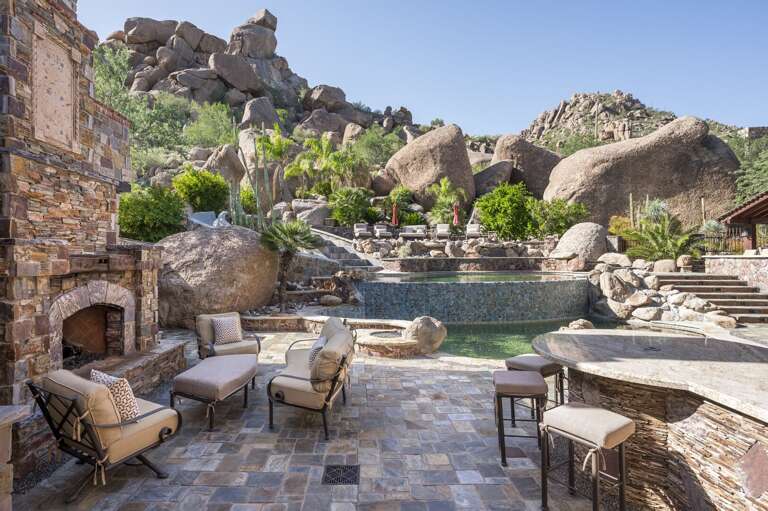 Experience a private desert oasis featuring a stunning multi-level pool, waterslide, outdoor fireplace, and luxurious lounge areas, all surrounded by breathtaking natural rock formations.
