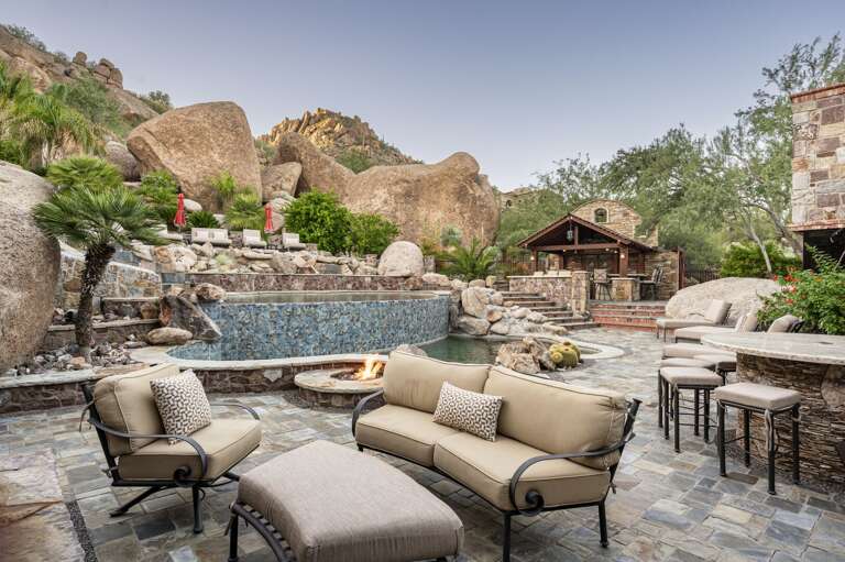 Experience a private desert oasis featuring a stunning multi-level pool, waterslide, outdoor fireplace, and luxurious lounge areas, all surrounded by breathtaking natural rock formations.