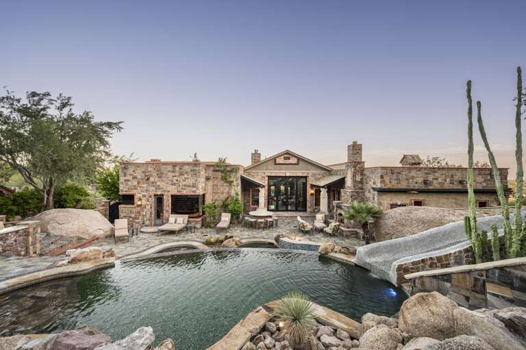 Experience a private desert oasis featuring a stunning multi-level pool, waterslide, outdoor fireplace, and luxurious lounge areas, all surrounded by breathtaking natural rock formations.