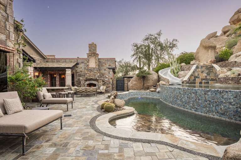Experience a private desert oasis featuring a stunning multi-level pool, waterslide, outdoor fireplace, and luxurious lounge areas, all surrounded by breathtaking natural rock formations.